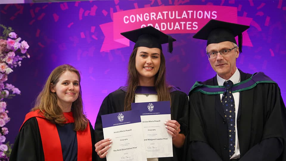 Jessica standing in her graduation gown and hat holding the award with a sign that says Congratulations graduate in Loughborough colours.